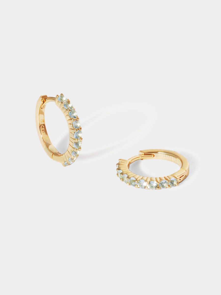 A pair of 9 karat gold huggies with 9 light blue aquamarine gemstones per huggie. Set on an architectural grey background with elongated shadows