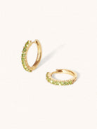 A pair of 9 karat gold huggies with 9 green peridot gemstones per huggie. Set on a cream background with elongated shadows