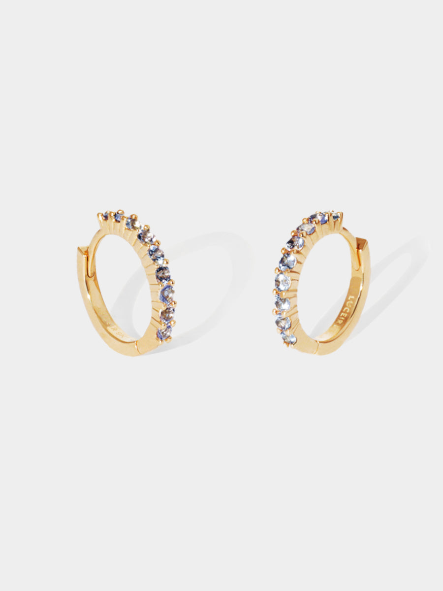 A pair of 9 karat gold huggies with 9 purple tanzanite stones per huggie. Set on an architectural grey background with elongated huggie shadows