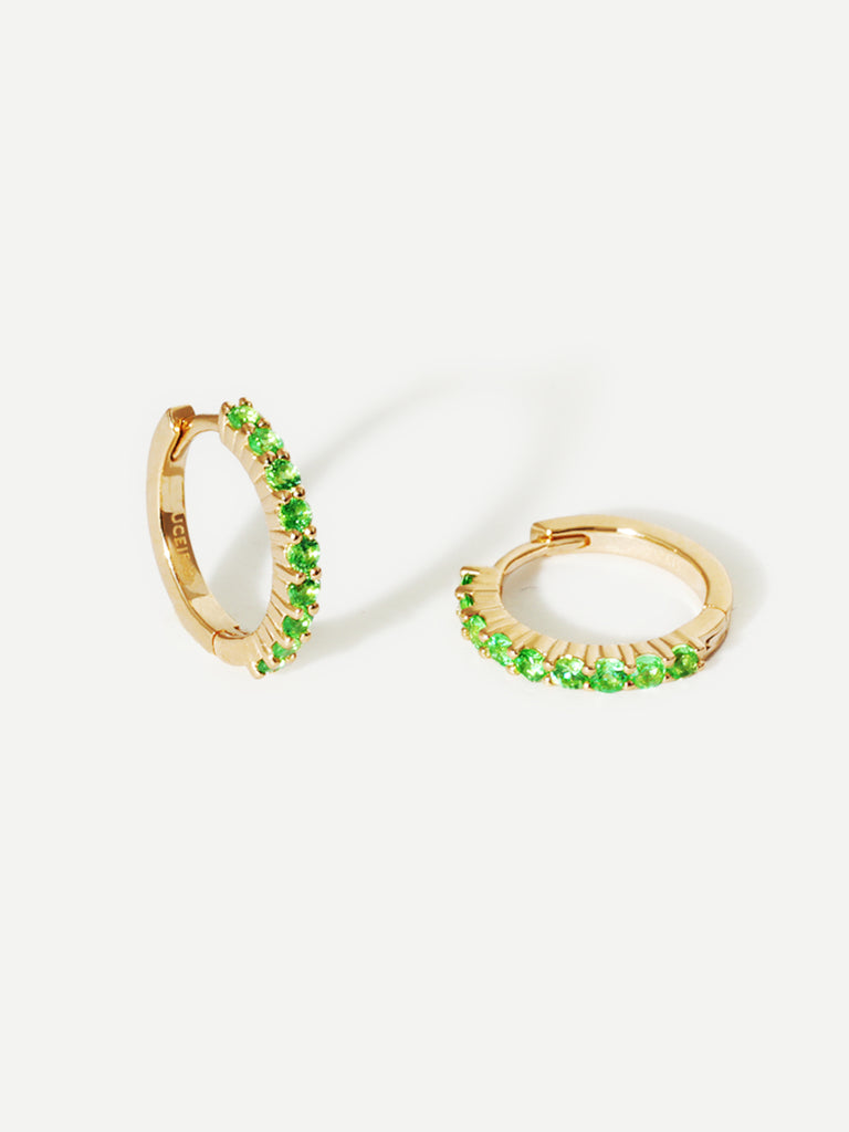 A pair of 9 karat gold huggies with 9 vibrant green tsavourite gemstones per huggie. Set on an architectural grey background with elongated shadows