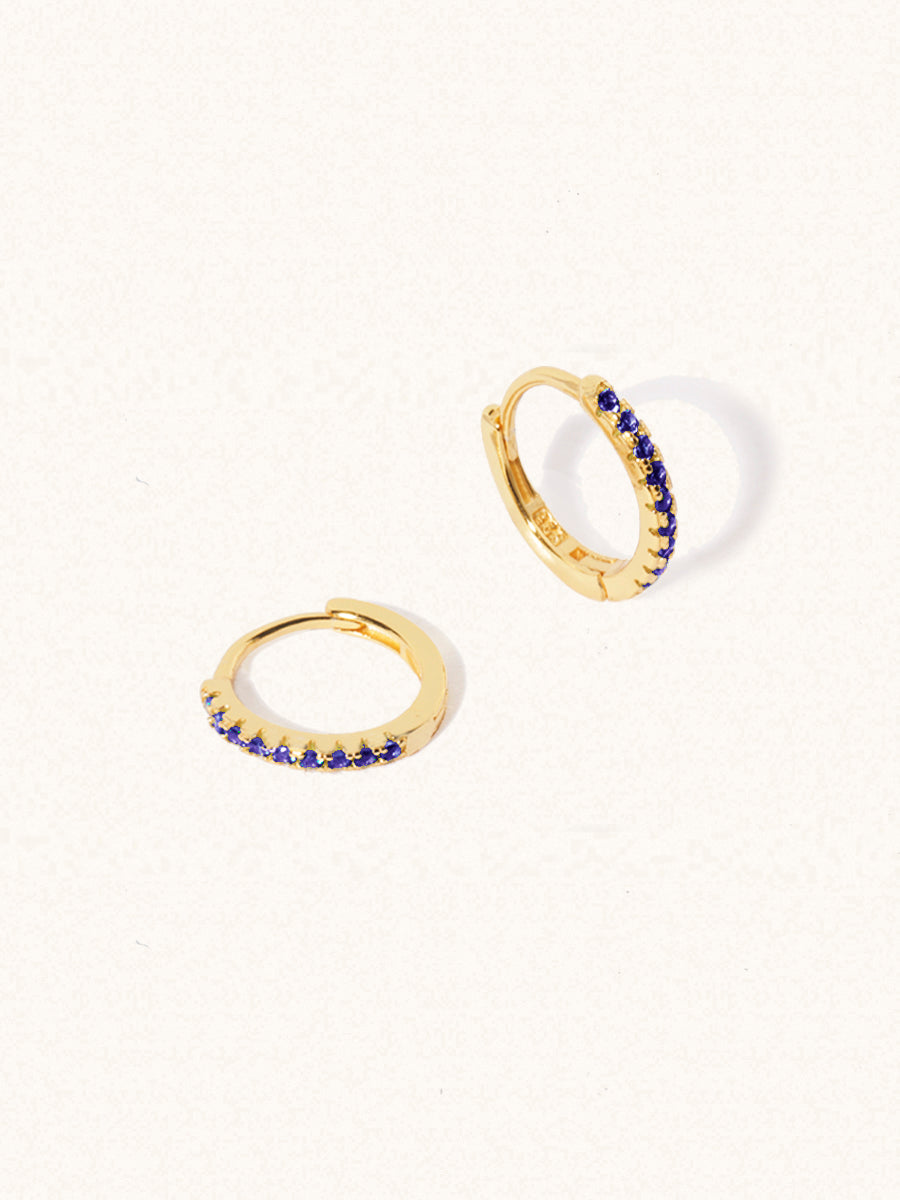 A pair of gold plated huggies with 10 deep blue quartz gemstones per huggie. Set on a cream background with elongated shadows