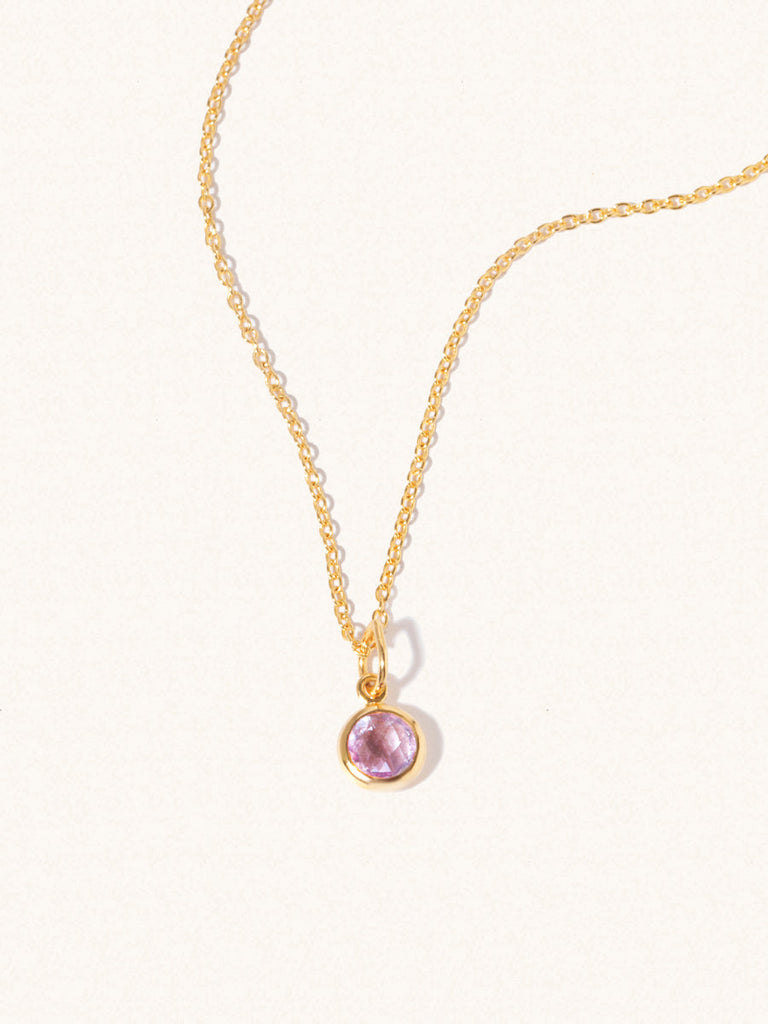 A gold delicate chain nacklace with an 8mm purple Brazilian Amethyst gemstone set in a round pendant. Set on an cream background with gentle shadowing.