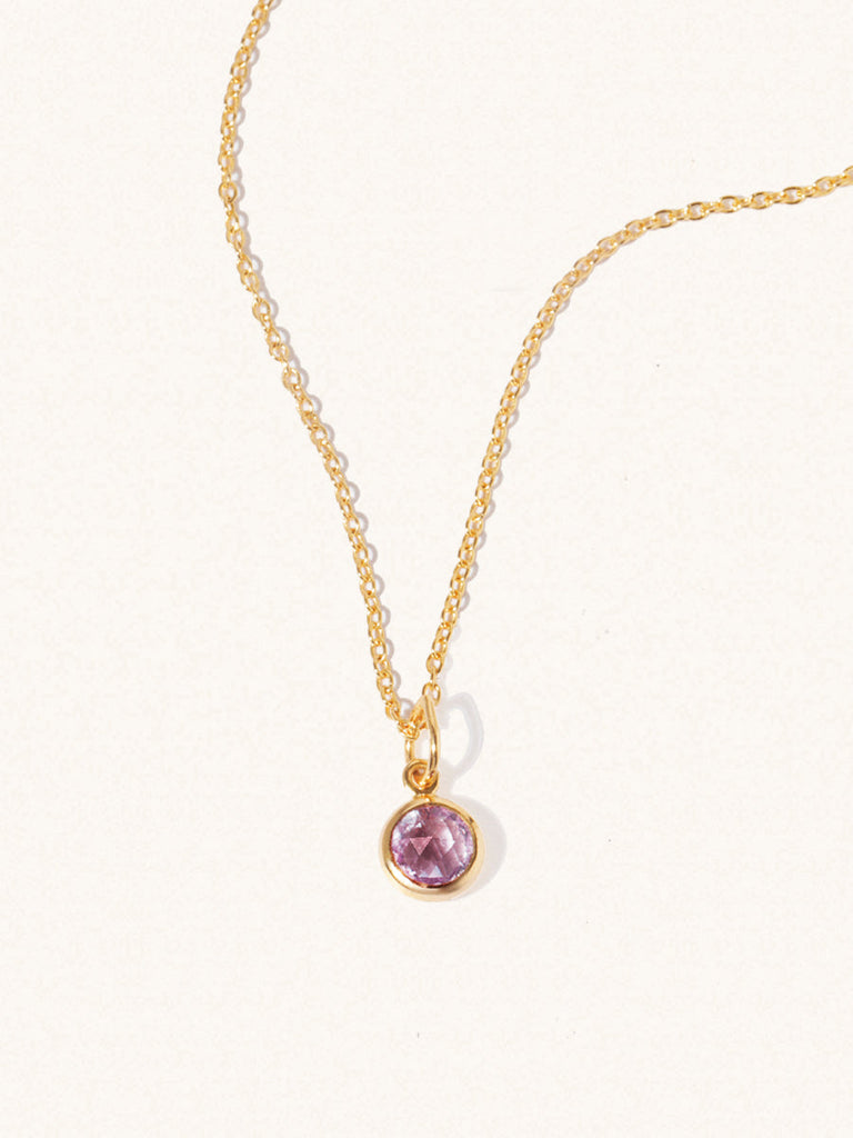 A gold delicate chain nacklace with an 8mm purple Brazilian Amethyst gemstone set in a round pendant. Set on an cream background with gentle shadowing.