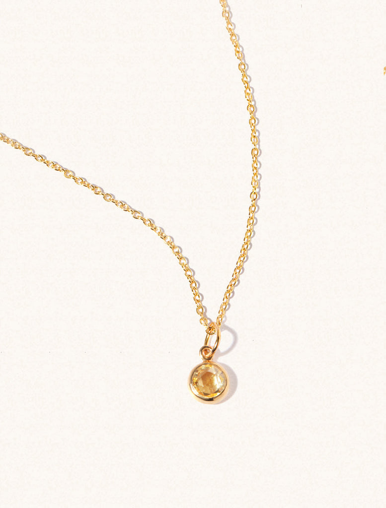 A gold delicate chain nacklace with an 8mm yellow citrine gemstone set in a round pendant. Set on an cream background with gentle shadowing.