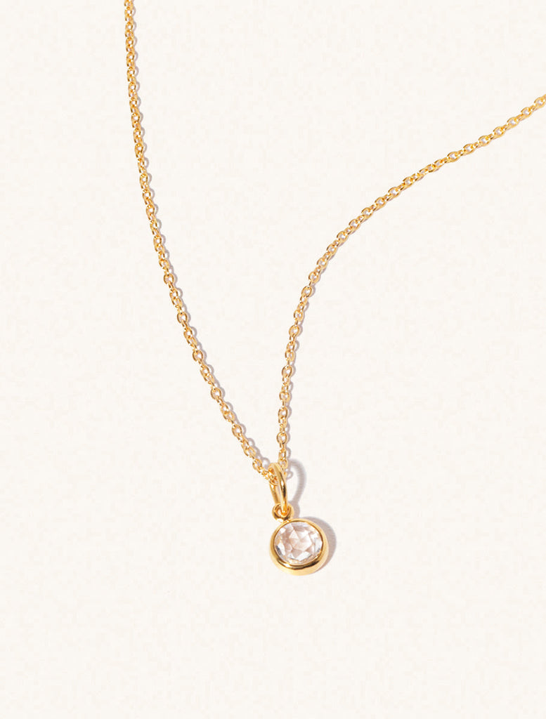 A gold delicate chain nacklace with an 8mm white crystal gemstone set in a round pendant. Set on an cream background with gentle shadowing.