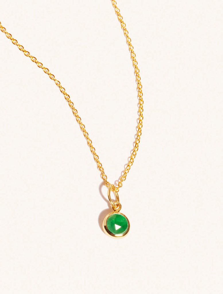 A gold delicate chain nacklace with an 8mm emerald quartz gemstone set in a round pendant. Set on an cream background with gentle shadowing.