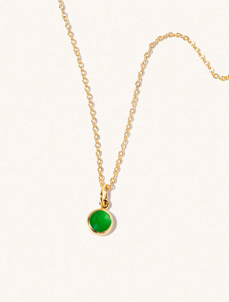 A gold delicate chain nacklace with an 8mm emerald quartz gemstone set in a round pendant. Set on an cream background with gentle shadowing.