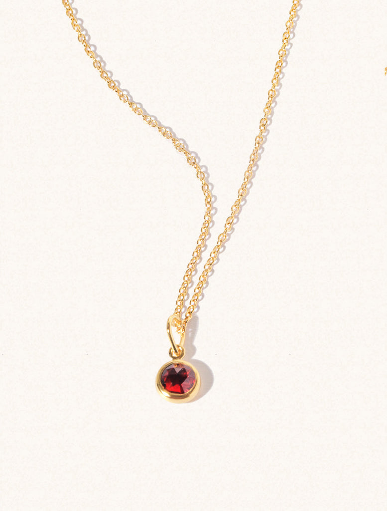 A gold delicate chain nacklace with an 8mm deep red Garnet gemstone set in a round pendant. Set on an cream background with gentle shadowing.