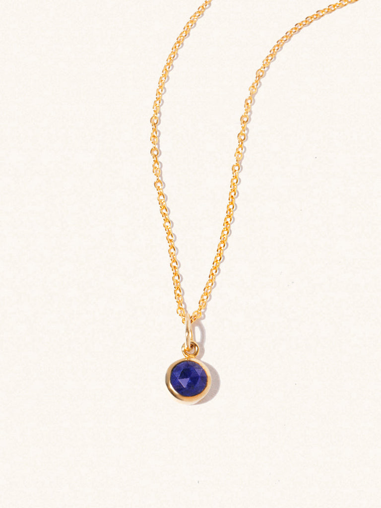 A gold delicate chain nacklace with an 8mm blue lapis lazuli gemstone set in a round pendant. Set on an cream background with gentle shadowing.