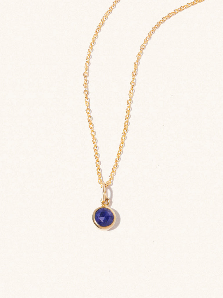 A gold delicate chain nacklace with an 8mm blue lapis lazuli gemstone set in a round pendant. Set on an cream background with gentle shadowing.