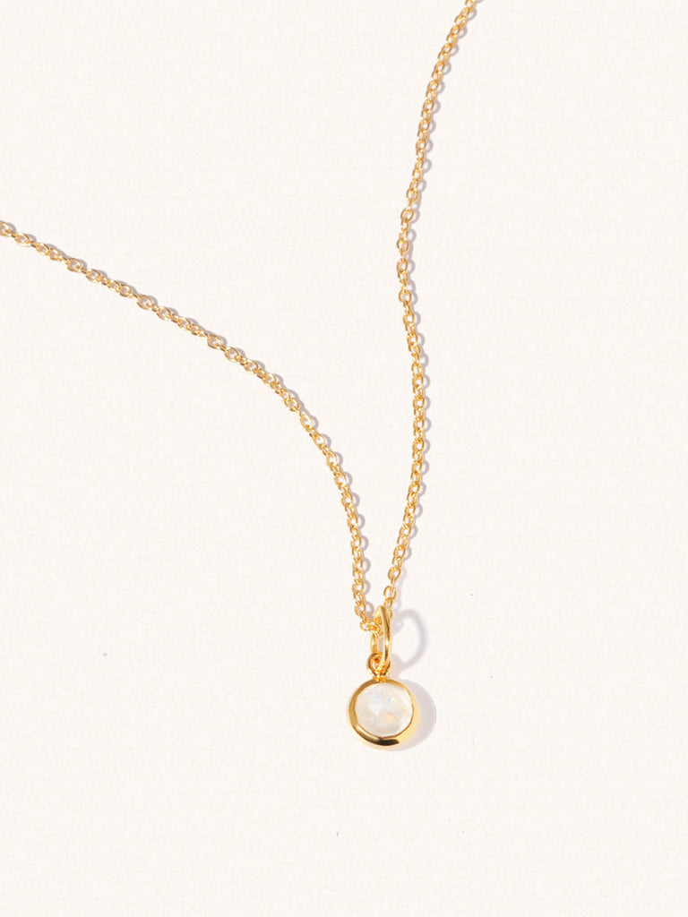 A gold delicate chain nacklace with an 8mm moonstone gemstone set in a round pendant. Set on an cream background with gentle shadowing.