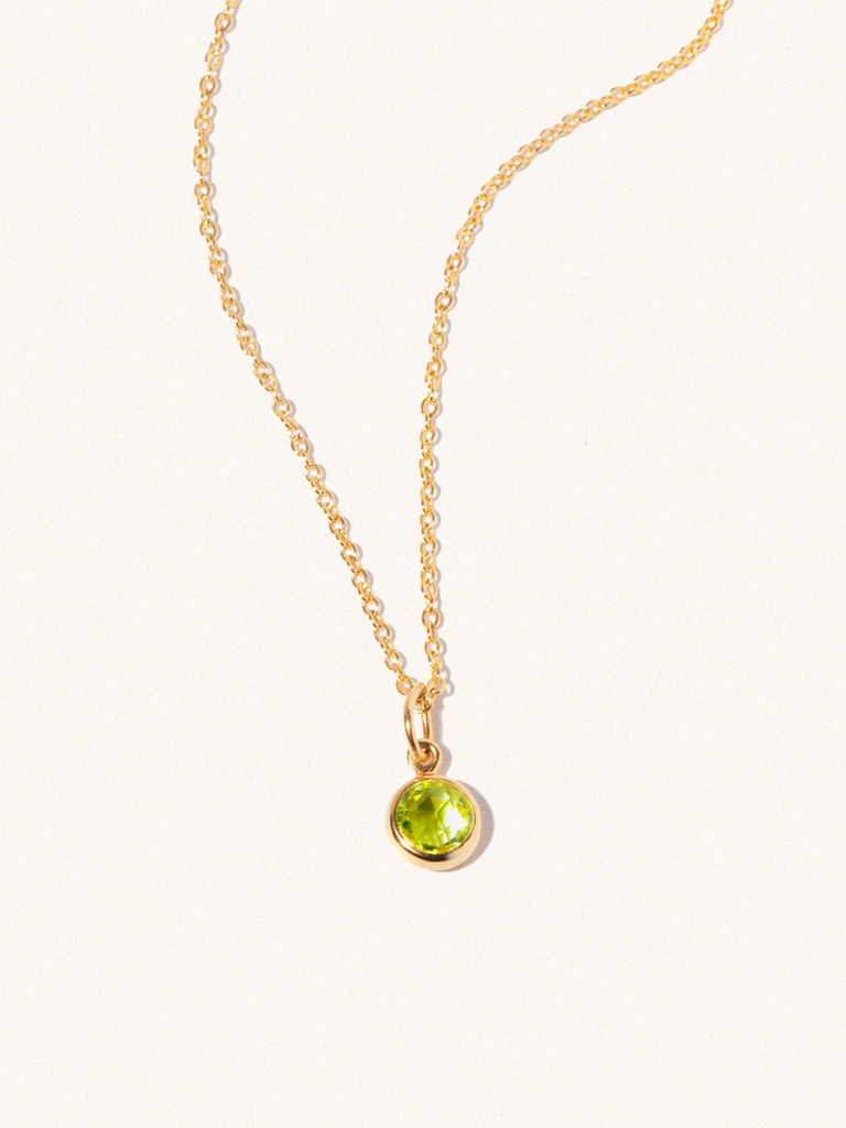 A gold delicate chain nacklace with an 8mm light green Peridot gemstone set in a round pendant. Set on an cream background with gentle shadowing.