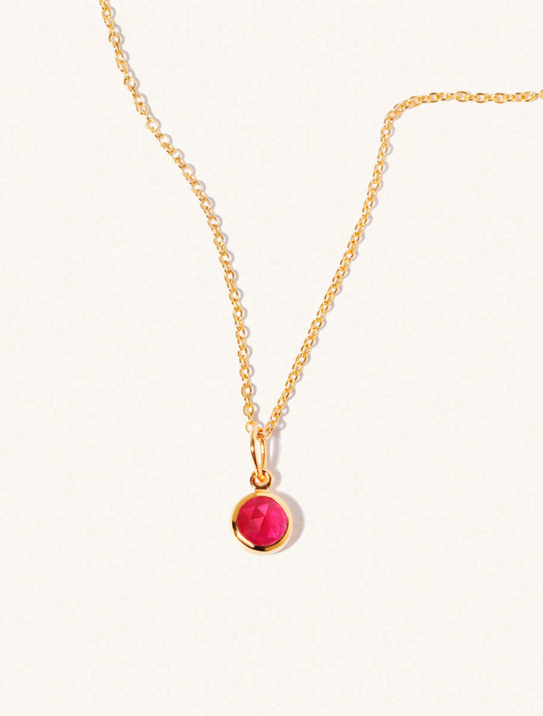 A gold delicate chain nacklace with an 8mm ruby quartz gemstone set in a round pendant. Set on an cream background with gentle shadowing.