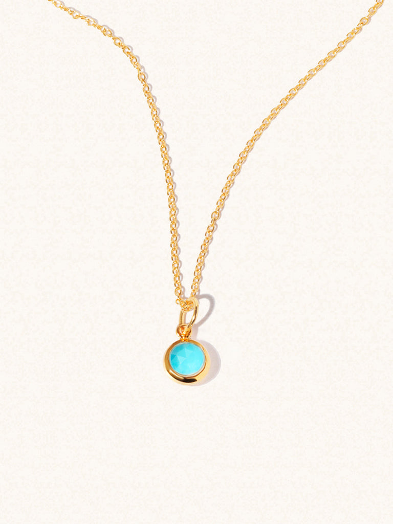 A gold delicate chain nacklace with an 8mm turquoise gemstone set in a round pendant. Set on an cream background with gentle shadowing.