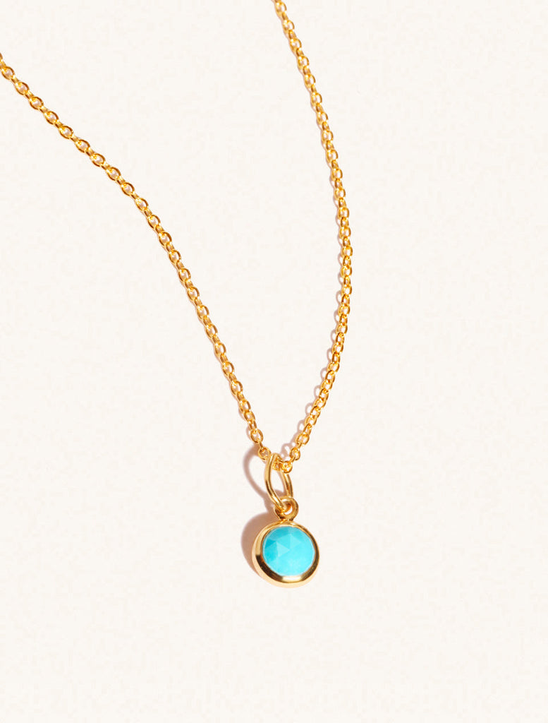 A gold delicate chain nacklace with an 8mm turquoise gemstone set in a round pendant. Set on an cream background with gentle shadowing.