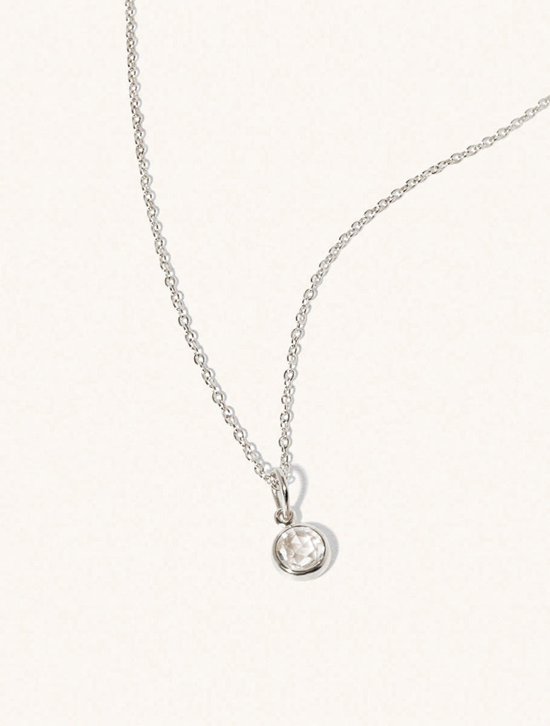 A sterling silver delicate chain nacklace with an 8mm white crystal gemstone set in a round pendant. Set on an cream background with gentle shadowing.