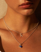 Close up shot of model neck and upper chest wearing two sterling silver necklaces. One with a letter A charm and the other a lower hanging blue lapis lazuli stone pendant in a circular setting