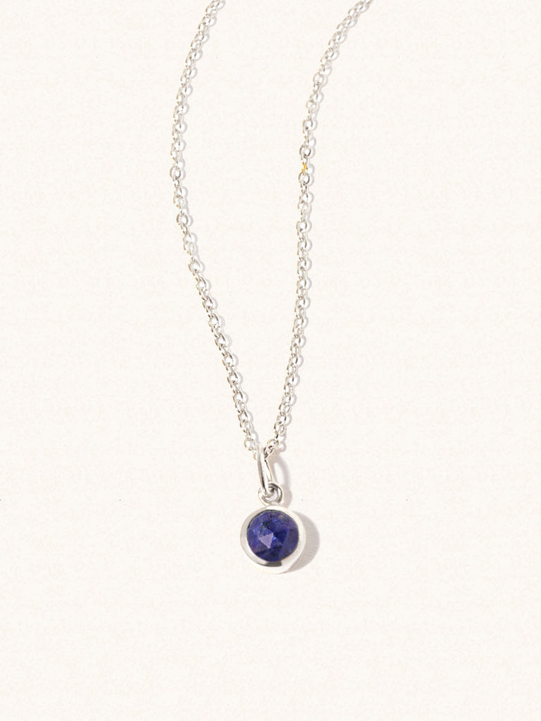 A sterling silver delicate chain nacklace with an 8mm blue lapis lazuli gemstone set in a round pendant. Set on an cream background with gentle shadowing.