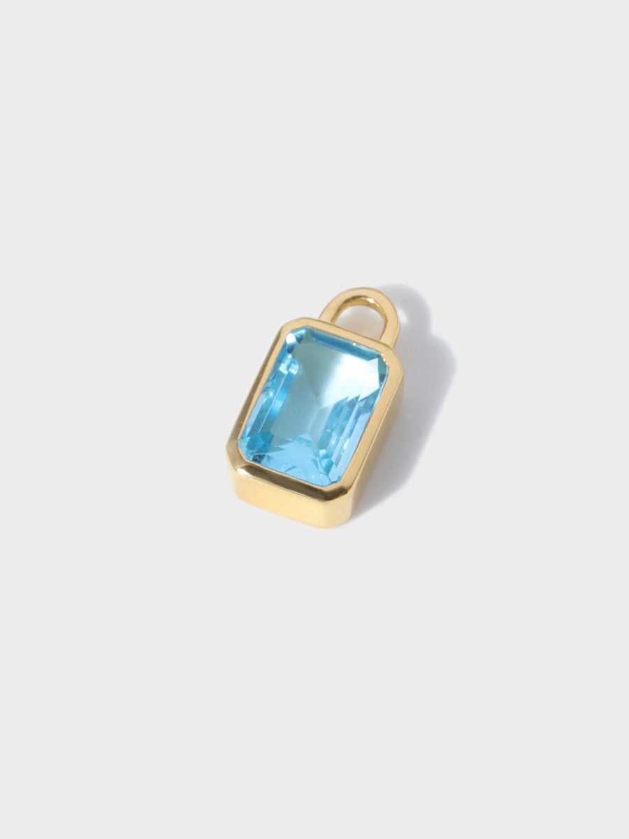 A large blue topaz baguette shaped charm set in a gold casing with large connection hoop at the top. Grey background with shadow