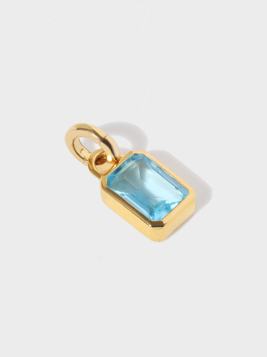 A large blue topaz baguette shaped charm set in a gold casing with large connection hoop at the top and clicker connector attahced. Grey background with shadow