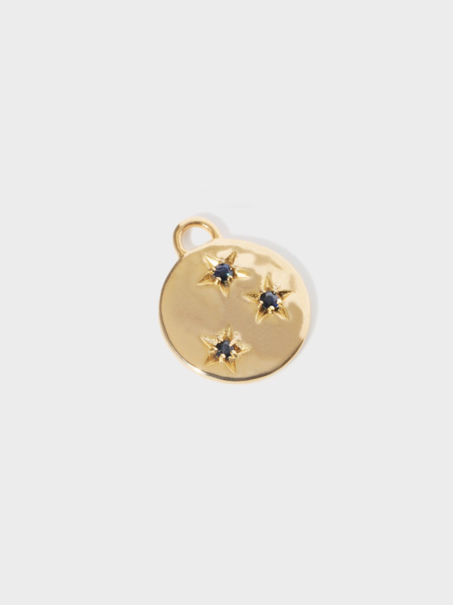 A gold circular necklace pendant set with 3 blue sapphire gemstones in star cutouts with a large hoop at the top. Grey background with shadow.