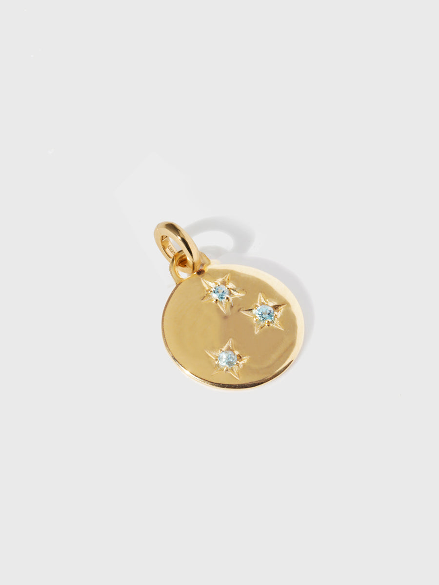 A gold circular necklace pendant set with 3 blue topaz gemstones in star cutouts with a large hoop at the top and clicker connector attached. Grey background with shadow.