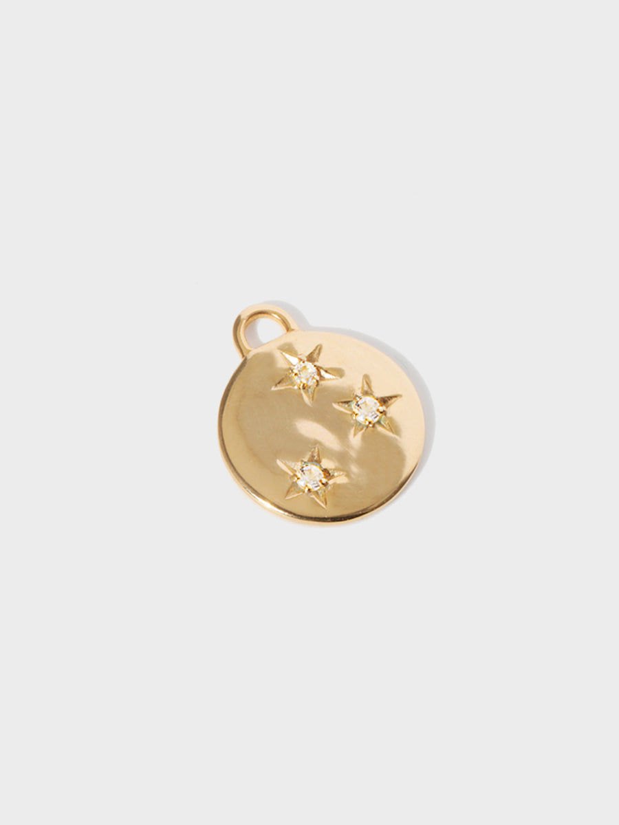 A gold circular necklace pendant set with 3 white topaz gemstones in star cutouts with a large hoop at the top. Grey background with shadow.