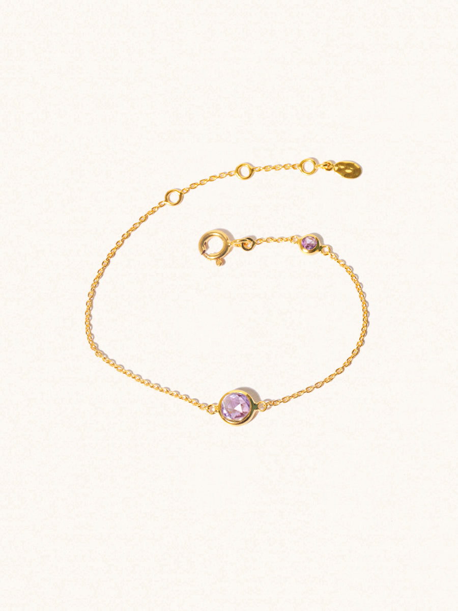 Delicate gold bracelet with two purple Brazilian Amethyst gemstones. One larger central 5mm gemstone and one smaller 2mm gemstone towards the round spring fastening. Set on a cream background with slight shadow.