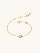 Delicate gold bracelet with two blue topaz gemstones. One larger central 5mm gemstone and one smaller 2mm gemstone towards the round spring fastening. Set on a cream background with slight shadow.