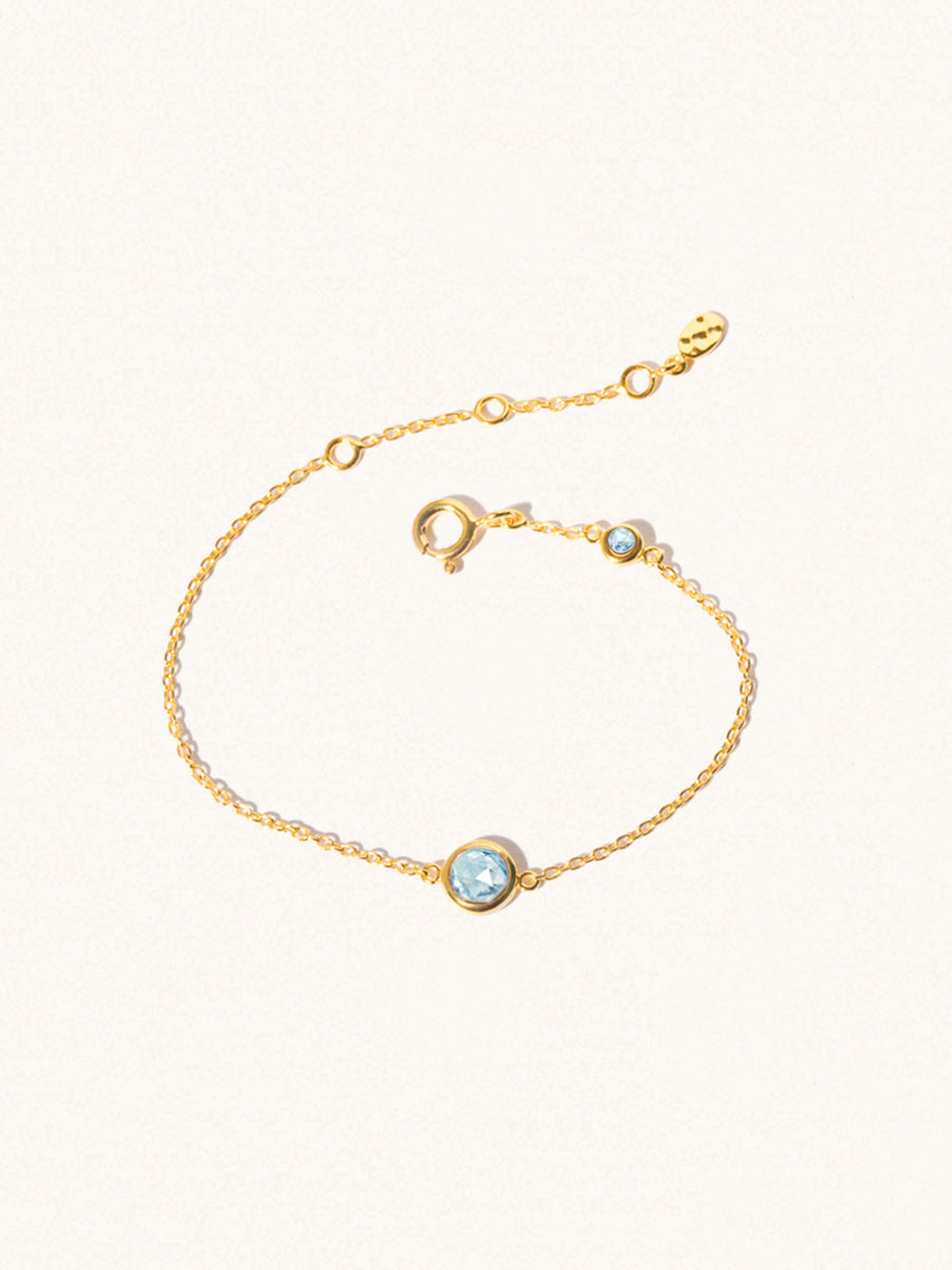 Delicate gold bracelet with two blue topaz gemstones. One larger central 5mm gemstone and one smaller 2mm gemstone towards the round spring fastening. Set on a cream background with slight shadow.