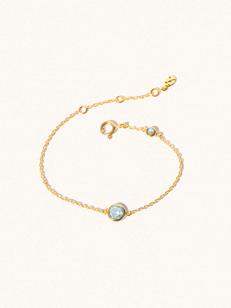 Delicate gold bracelet with two blue topaz gemstones. One larger central 5mm gemstone and one smaller 2mm gemstone towards the round spring fastening. Set on a cream background with slight shadow.