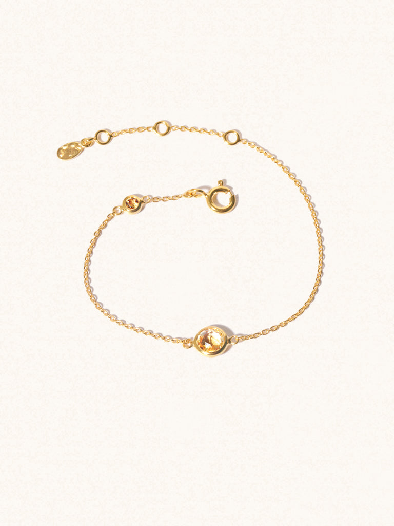 Delicate gold bracelet with two yellow citrine gemstones. One larger central 5mm gemstone and one smaller 2mm gemstone towards the round spring fastening. Set on a cream background with slight shadow.