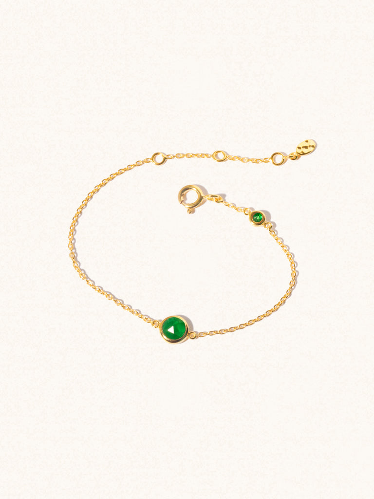 Delicate gold bracelet with two emerald quartz gemstones. One larger central 5mm gemstone and one smaller 2mm gemstone towards the round spring fastening. Set on a cream background with slight shadow.