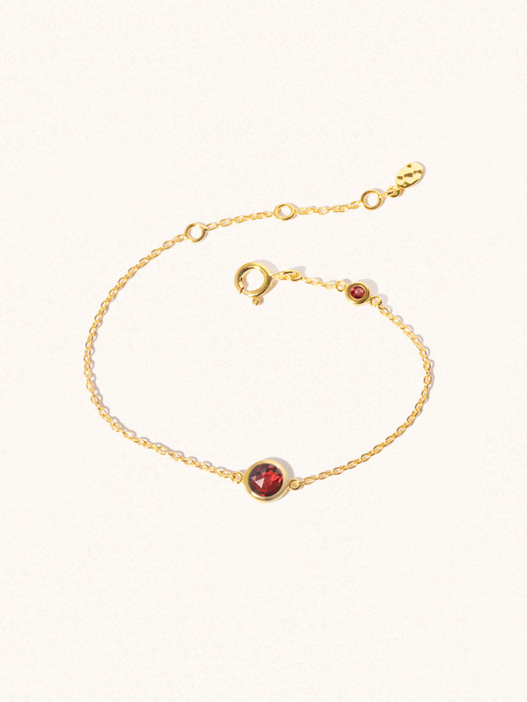 Delicate gold bracelet with two deep red Garnet gemstones. One larger central 5mm gemstone and one smaller 2mm gemstone towards the round spring fastening. Set on a cream background with slight shadow.