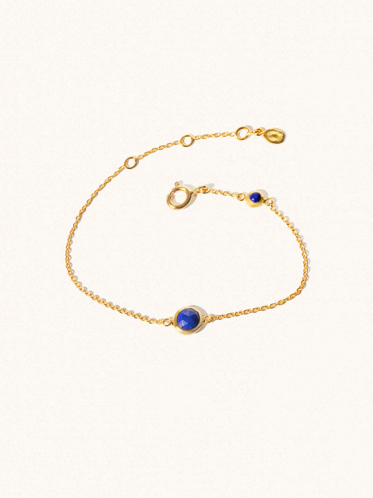 Delicate gold bracelet with two blue lapiz lazuli gemstones. One larger central 5mm gemstone and one smaller 2mm gemstone towards the round spring fastening. Set on a cream background with slight shadow.