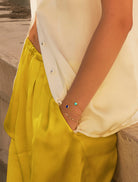 A cropped shot of a woman wearing mustard silk trousers and crea silk blouse wearing two gold birthstone bracelets with turquoise and lapis lazuli gemstones as she casually has her left hand in her trouser pocket whilst leaning against a waist height stone wall.