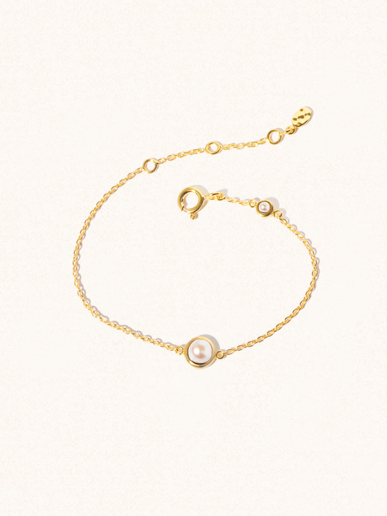 Delicate gold bracelet with two mother of pearl gemstones. One larger central 5mm pearl gemstone and one smaller 2mm pearl gemstone towards the round spring fastening. Set on a cream background with slight shadow.