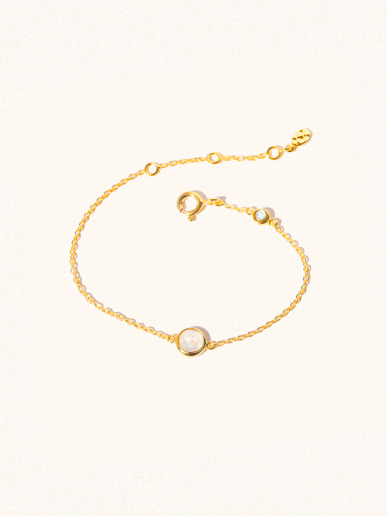 Delicate gold bracelet with two iridescent moonstone gemstones. One larger central 5mm moonstone gemstone and one smaller 2mm moonstone gemstone towards the round spring fastening. Set on a cream background with slight shadow.