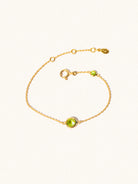 Delicate gold bracelet with two green peridot gemstones. One larger central 5mm gemstone and one smaller 2mm gemstone towards the round spring fastening. Set on a cream background with slight shadow.