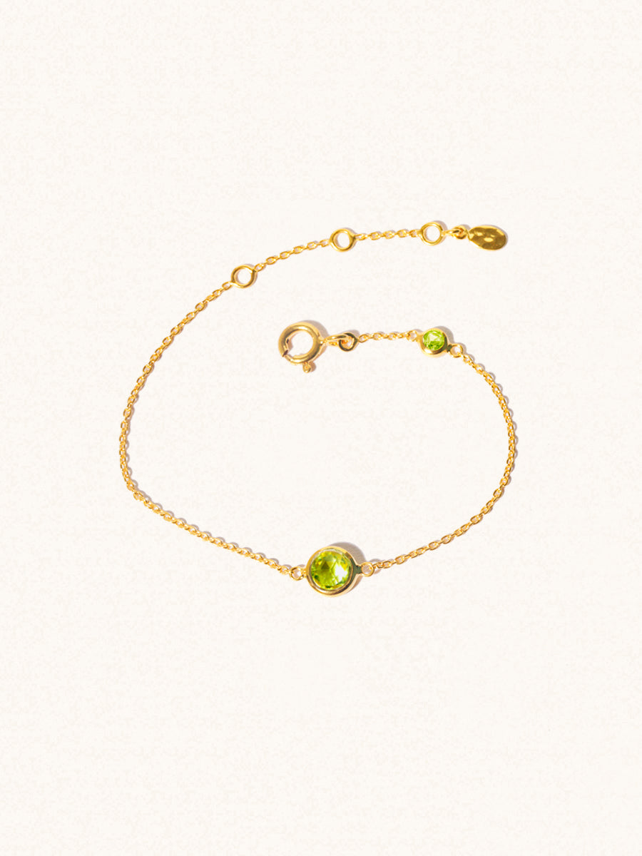 Delicate gold bracelet with two green peridot gemstones. One larger central 5mm gemstone and one smaller 2mm gemstone towards the round spring fastening. Set on a cream background with slight shadow.