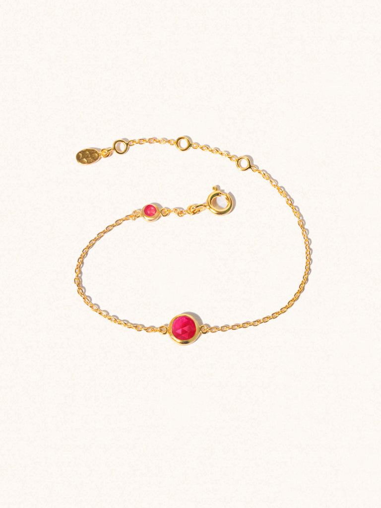 Delicate gold bracelet with two ruby quartz gemstones. One larger central 5mm gemstone and one smaller 2mm gemstone towards the round spring fastening. Set on a cream background with slight shadow.