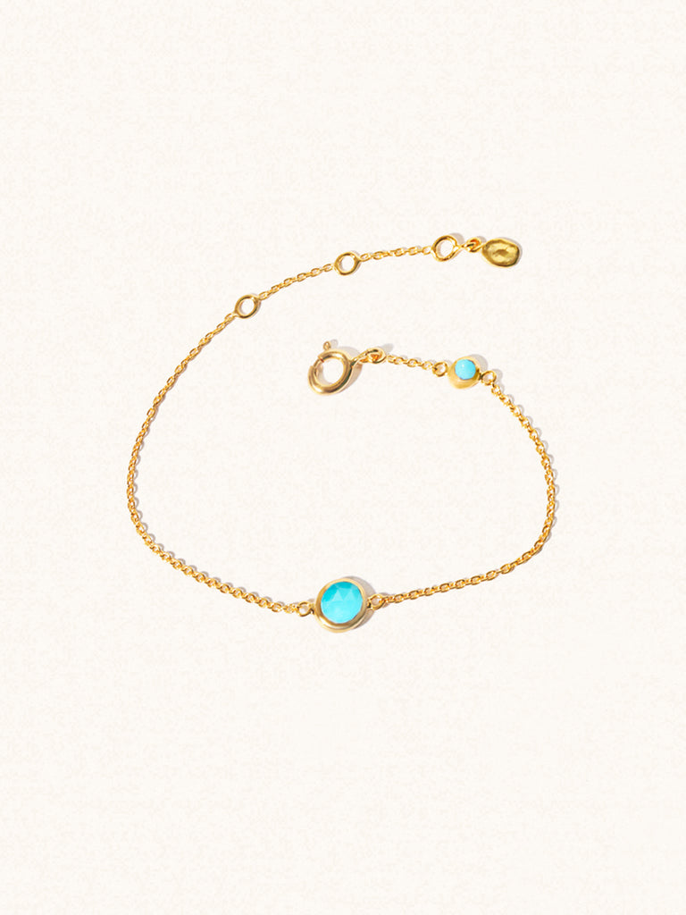 Delicate gold bracelet with two truquoise gemstones. One larger central 5mm gemstone and one smaller 2mm gemstone towards the round spring fastening. Set on a cream background with slight shadow.