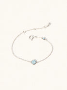 Delicate sterling silver bracelet with two blue topaz gemstones. One larger central 5mm gemstone and one smaller 2mm gemstone towards the round spring fastening. Set on a cream background with slight shadow.