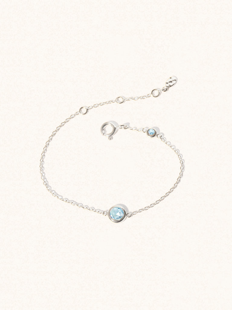 Delicate sterling silver bracelet with two blue topaz gemstones. One larger central 5mm gemstone and one smaller 2mm gemstone towards the round spring fastening. Set on a cream background with slight shadow.