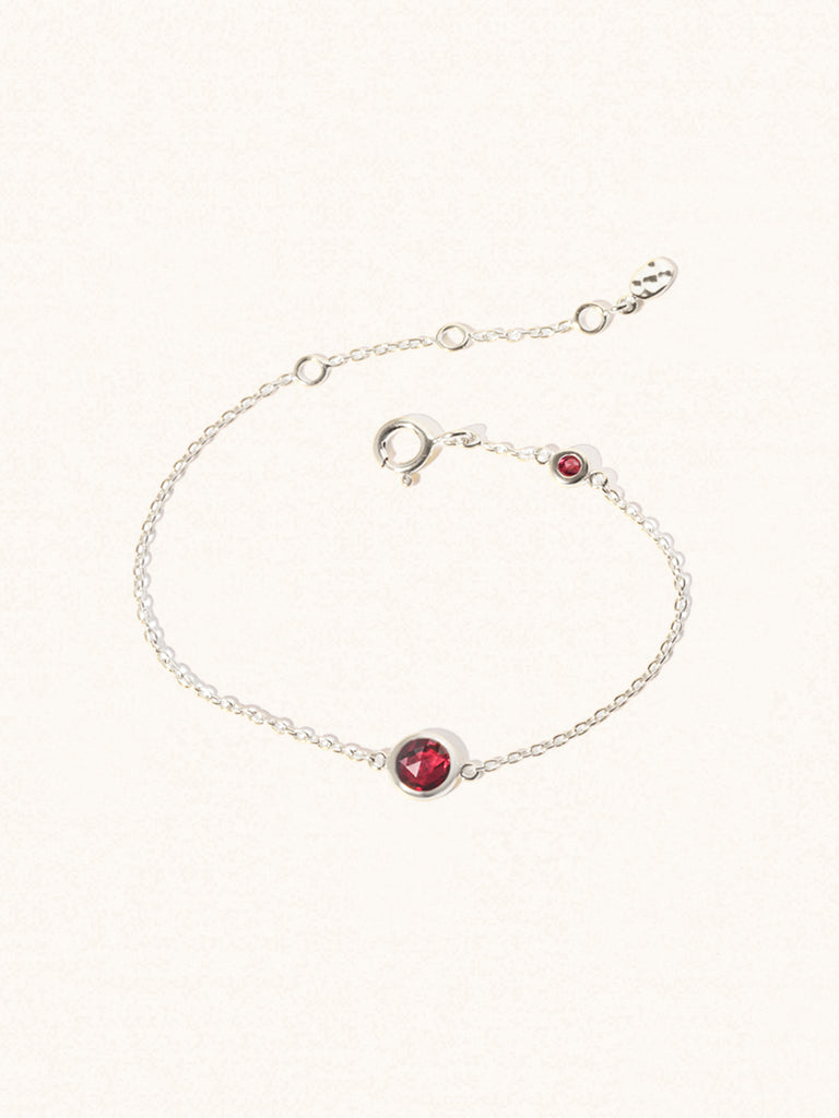 Delicate sterling silver bracelet with two deep red Garnet gemstones. One larger central 5mm gemstone and one smaller 2mm gemstone towards the round spring fastening. Set on a cream background with slight shadow.