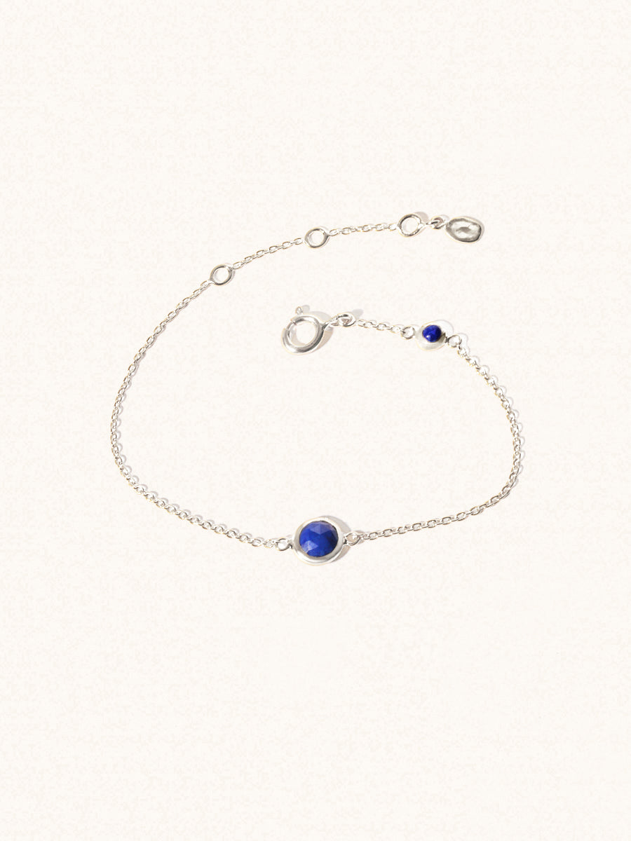 Delicate sterling silver bracelet with two blue lapiz lazuli gemstones. One larger 5mm central gemstone and one smaller 2mm gemstone towards the round spring fastening. Set on a cream background with slight shadow.