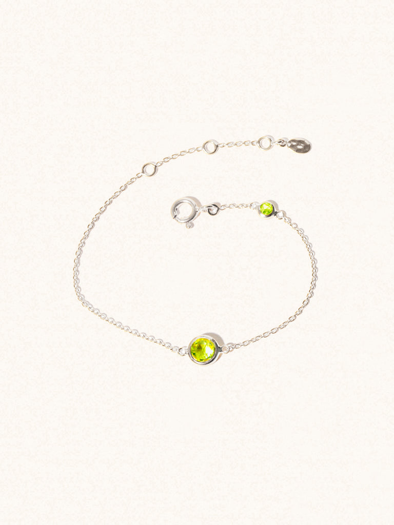 Delicate sterling silver bracelet with two green peridot gemstones. One larger 5mm central gemstone and one smaller 2mm gemstone towards the round spring fastening. Set on a cream background with slight shadow.