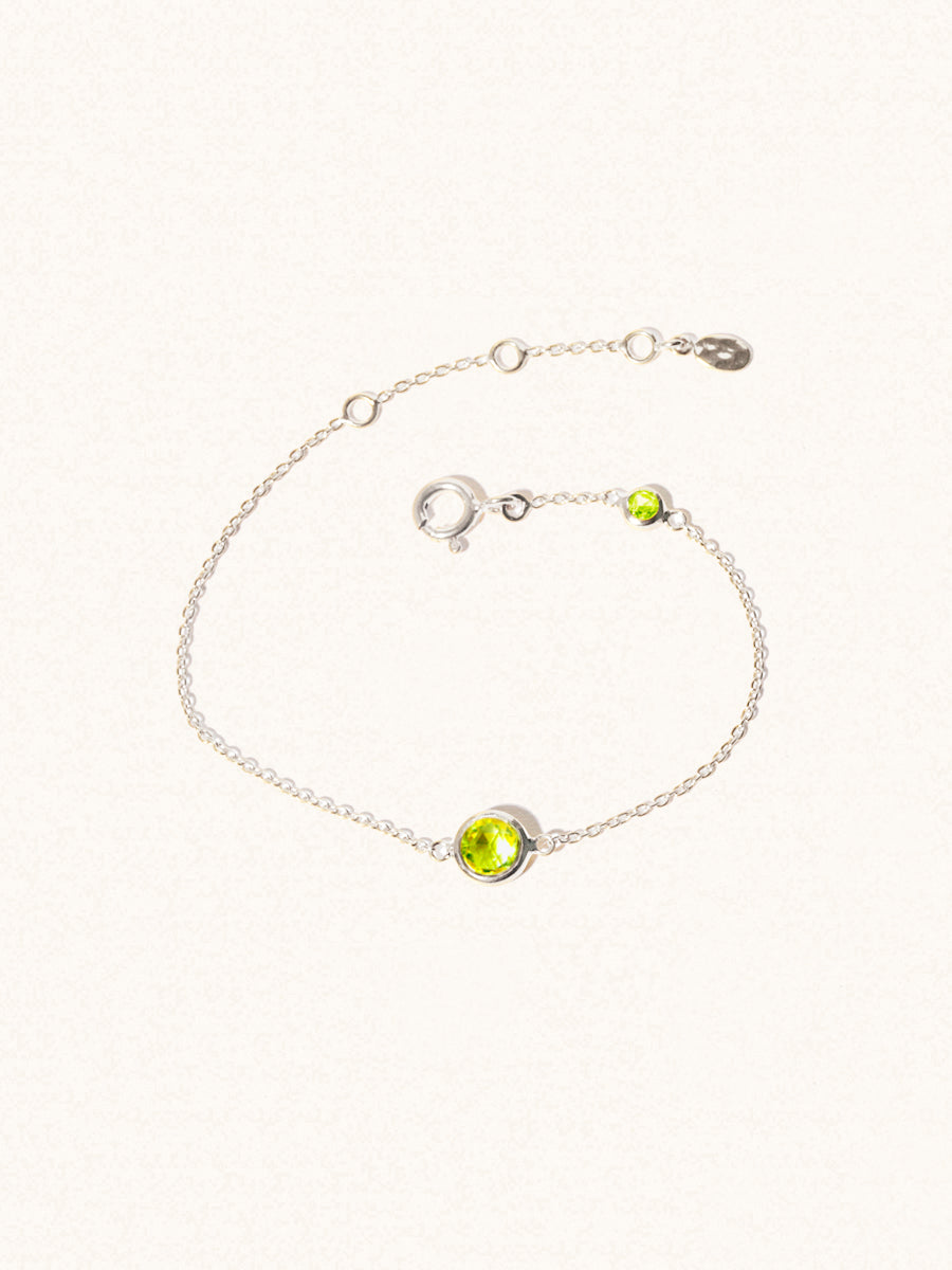 Delicate sterling silver bracelet with two green peridot gemstones. One larger 5mm central gemstone and one smaller 2mm gemstone towards the round spring fastening. Set on a cream background with slight shadow.