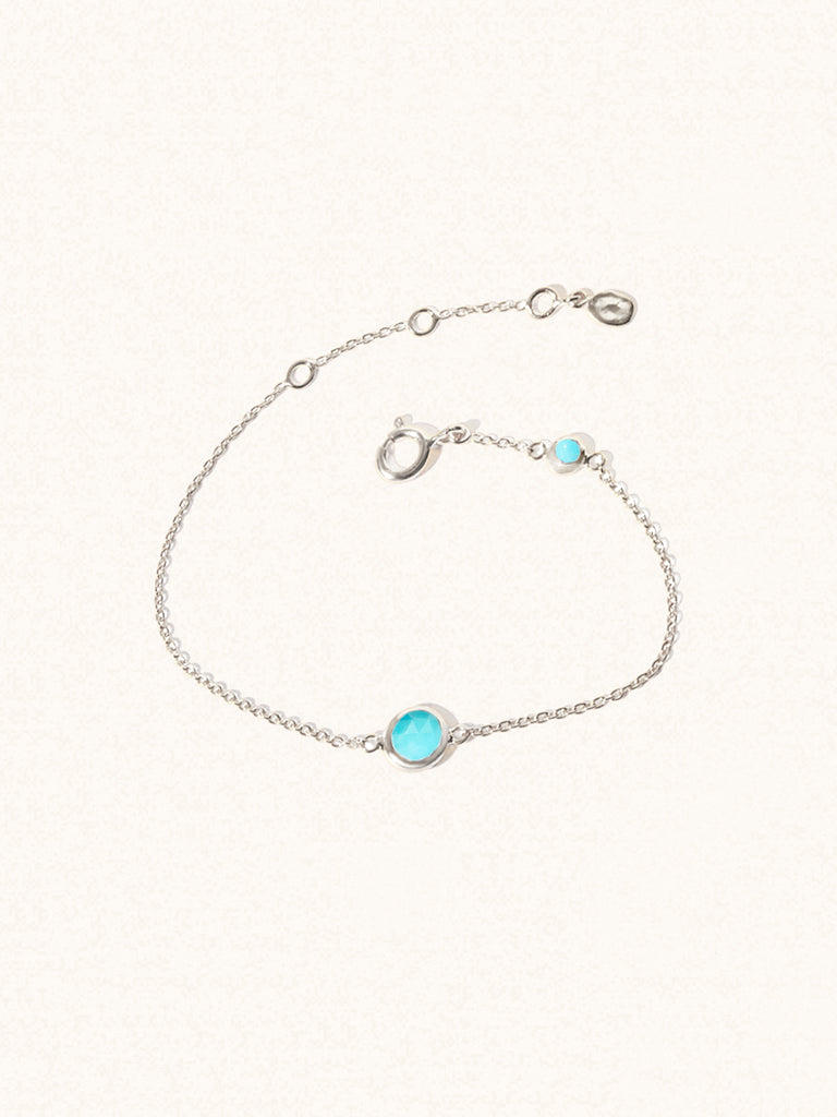 Delicate sterling silver bracelet with two truquoise gemstones. One larger central 5mm gemstone and one smaller 2mm gemstone towards the round spring fastening. Set on a cream background with slight shadow.