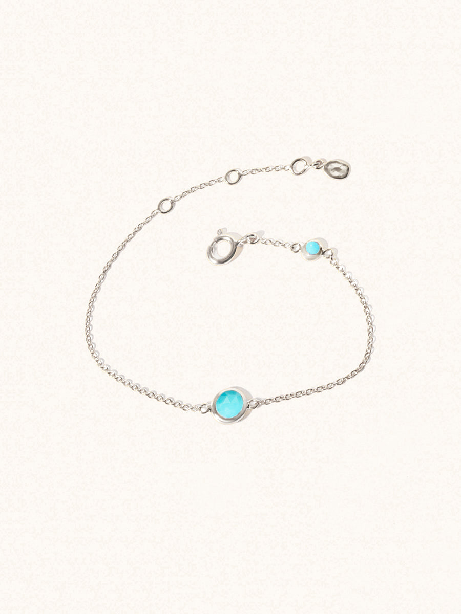 Delicate sterling silver bracelet with two truquoise gemstones. One larger central 5mm gemstone and one smaller 2mm gemstone towards the round spring fastening. Set on a cream background with slight shadow.